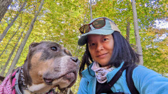 a woman wearing a baseball cap, jacket and sunglasses on her head takes a selfie in a forest with a dog